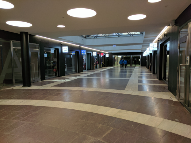 Langer Flur mit Menschen, die gehen, beleuchtet von Deckenlampen, flankiert von Säulen und Glastüren, mit Texttafeln, die an ein belebtes Rathaus erinnern.