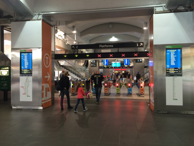 Menschen gehen durch einen Bahnhof mit Gepäck, Ticketautomat im Blick und Deckenbeleuchtung.
