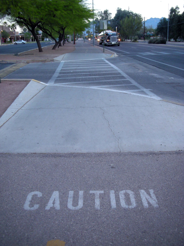 Stadtstraße mit fahrenden Fahrzeugen, ein Gehweg mit Warnschild in der Nähe eines Baumes, Strommasten, Straßenlaternen, Bauzäune, Grünfläche, Gebäude, entfernte Berge und einen klaren blauen Himmel.