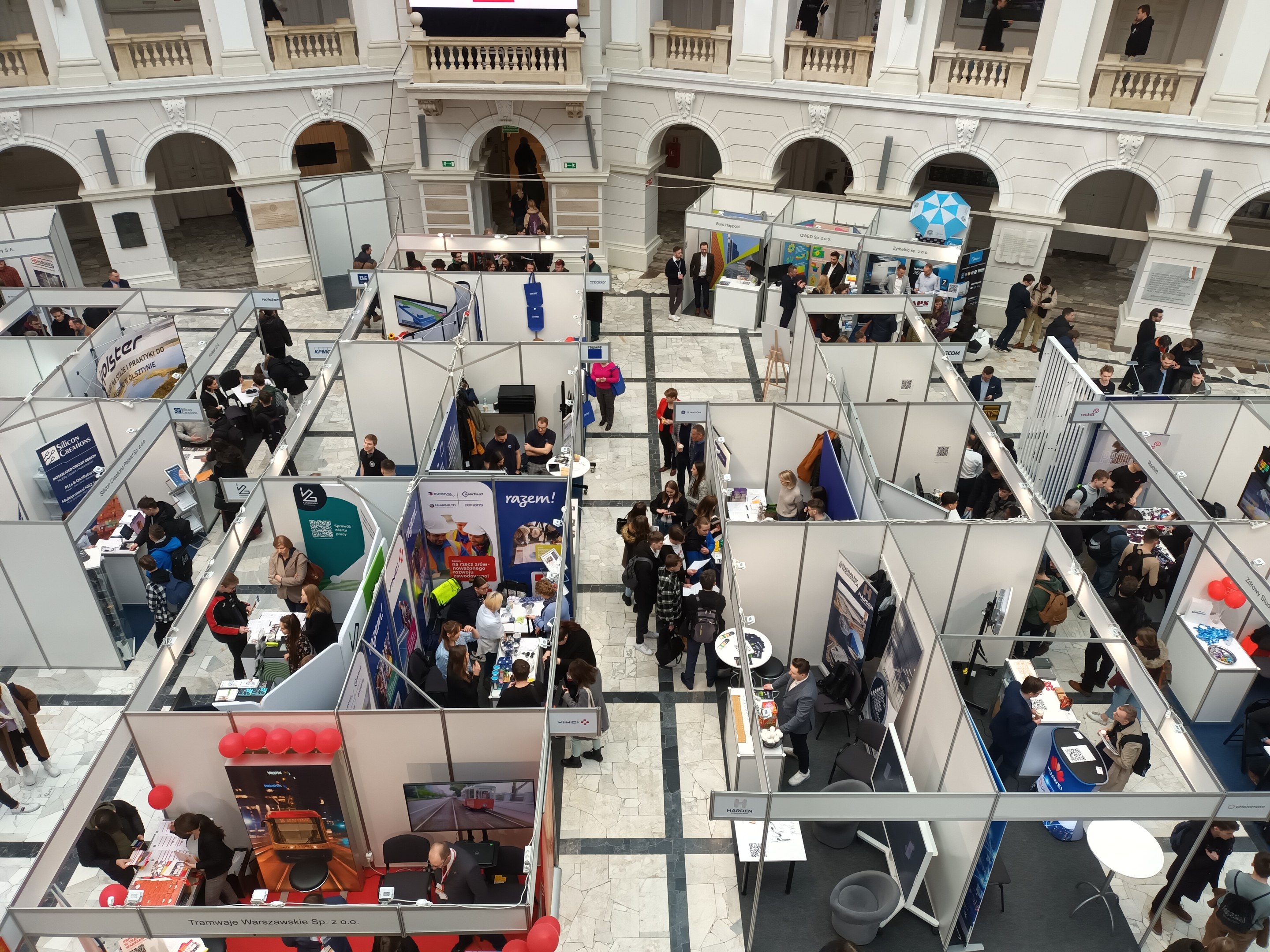 Große Gruppe von Menschen auf einer internationalen Messe mit Ständen, Bannern, Tischen, Stühlen, Luftballons und einem Gebäude mit Bögen, Säulen, Geländern und einem Bildschirm im Hintergrund.