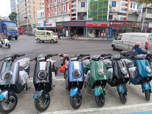 Eine Reihe von Elektro-Scootern, die auf einer Straße geparkt sind, mit einer Person in einer Mütze davor, Gebäuden, Verkehrszeichen, Bäumen und einem klaren blauen Himmel im Hintergrund.
