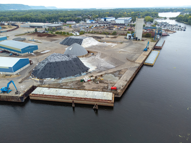 Luftaufnahme eines großen Gewässers mit einem zentralen Steg, umgeben von Gebäuden, Schuppen, Fahrzeugen, Kränen und anderen Strukturen, mit Bäumen, Hügeln und einem klaren blauen Himmel im Hintergrund.