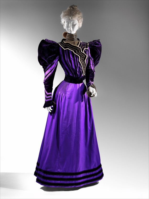 Eine Schaufensterpuppe trägt ein bodenlanges lila Abendkleid mit einem schwarzen und goldenen gemusterten Stoff, das eine enge Taille, einen weiten Rock, goldene Stickerei, Perlen und eine Spitzenverzierung an der Taille hat, kombiniert mit einem eleganten Hairstyle und zarten Ohrringen.