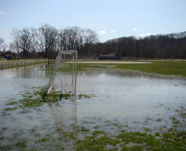 Ein Fußballtor steht in der Mitte eines überfluteten Feldes, umgeben von Gras, einem Zaun, Bäumen, Häusern und einem bewölkten Himmel.