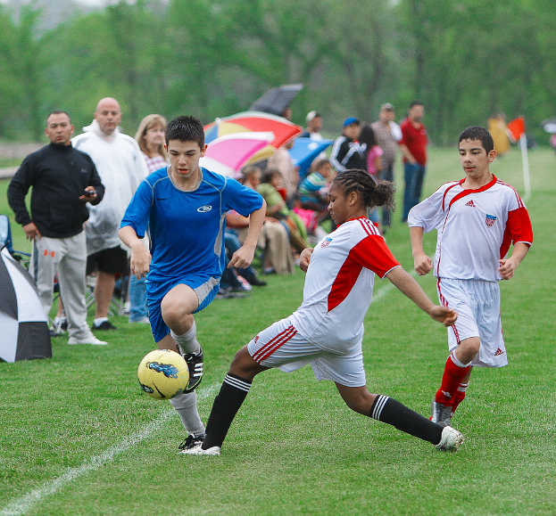 Drei Personen spielen Fußball auf einem Feld, hinter ihnen steht und sitzt eine Menge Menschen mit Schirmen, und im Hintergrund sind Bäume zu sehen.