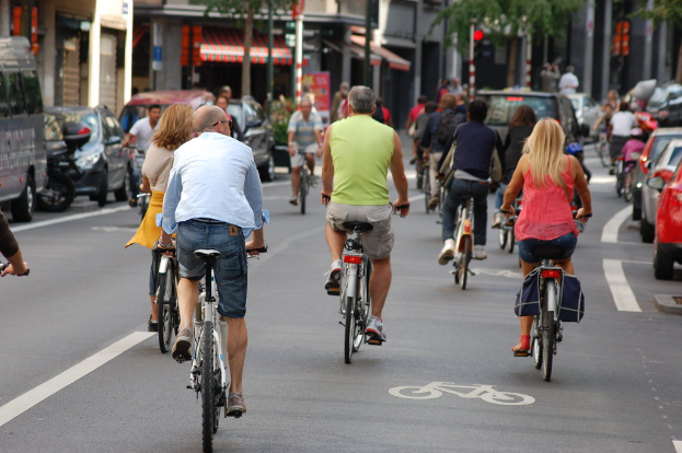 Radfahrer auf einer Straße mit Autos auf der linken und rechten Seite, mit Geschäften, einem Zelt, Bäumen und Gebäuden im Hintergrund.