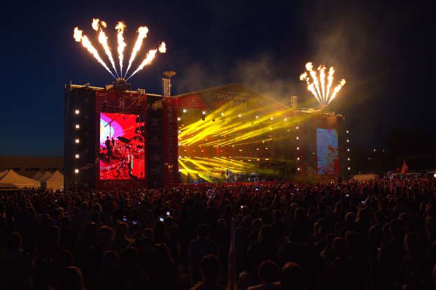 Eine große Menschenmenge auf einem Musikfestival mit einer Bühne in der Mitte, Musiker, die Instrumente unter hellen Scheinwerfern spielen und Banner, Zelte links und ein dunkler Himmel mit Feuerwerk im Hintergrund.