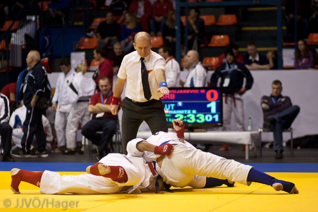 Zwei Judoka im Kampf mit einem Schiedsrichter, der zusieht, umgeben von sitzenden und stehenden Zuschauern, Geländern, Treppen und einer Anzeigetafel im Hintergrund.