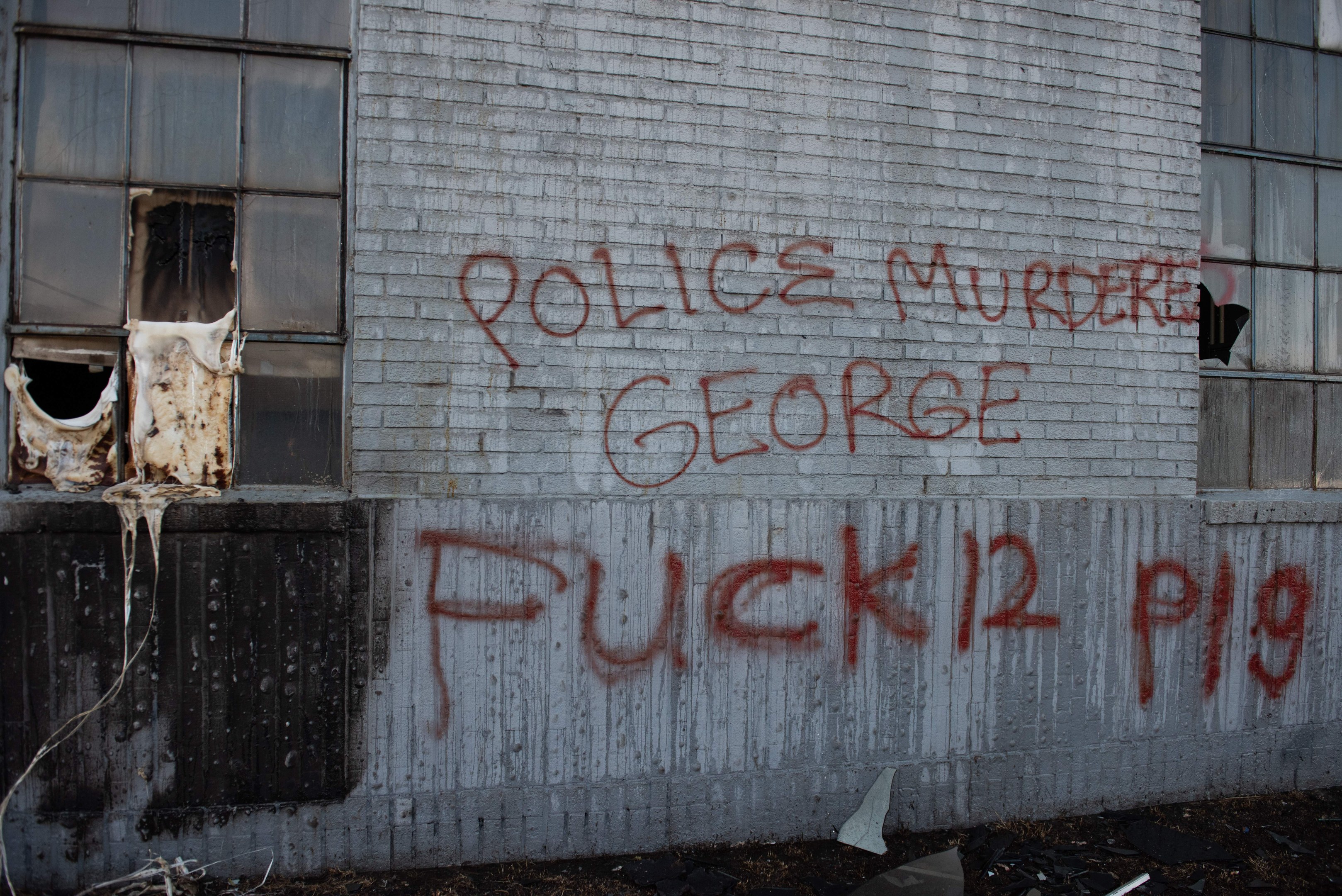 Graffiti an einer Gebäudewand mit der Aufschrift "Polizei ermordet George Fuck 12 P19" mit Fenstern und verstreuten Gegenständen davor.