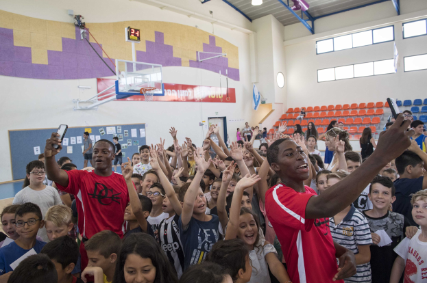 Kinder auf einem Basketballfeld mit Mobiltelefonen stehend, mit einer Anschlagtafel, Uhr, Torstange, Basketballkorb, Deckenleuchten, Stühlen und Fenstern im Hintergrund.