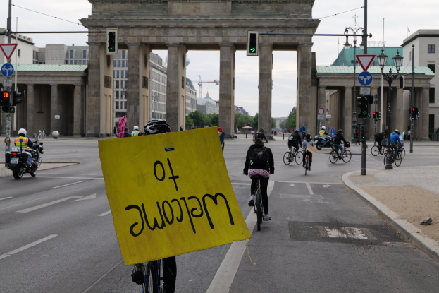Eine Gruppe von Menschen, die Fahrräder vor dem Brandenburger Tor in Berlin, Deutschland, fahren, wobei einer ein gelbes Schild hält und die Straße mit Laternen und Ampeln gesäumt ist.