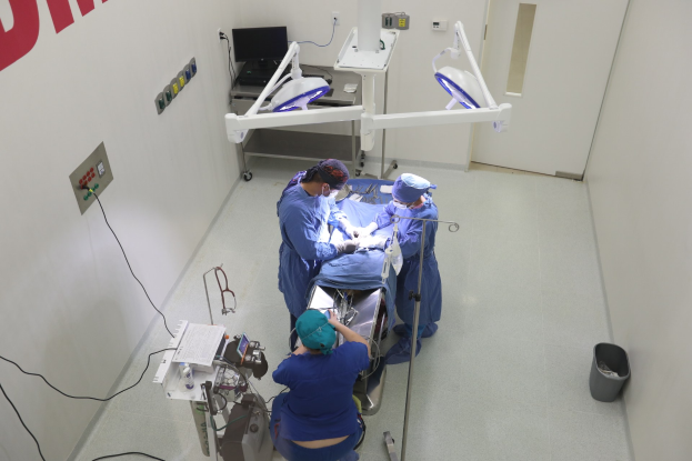 Eine Gruppe von Chirurgen führt einen Eingriff in einem Operationssaal durch, mit einem Patienten auf einer Trage umgeben von medizinischer Ausrüstung, Text an der linken Wand, einem Objekt auf dem Boden rechts und verschiedenen medizinischen Geräten im Hintergrund.