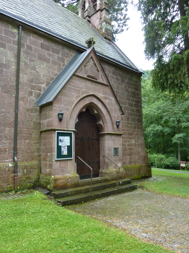 Eine alte steinerne Kirche mit einem Turm und einem Glockenturm, einer Tür, Treppen mit Geländern, einer Tafel mit Text, einem Rohr, Gras, Bäumen im Hintergrund und einem klaren blauen Himmel.