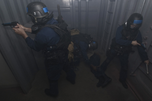 Eine Gruppe von Polizisten in Einsatzausrüstung, einer mit einer Waffe, neben einem Metallbehälter und einer Leiter in einem begrenzten Raum.