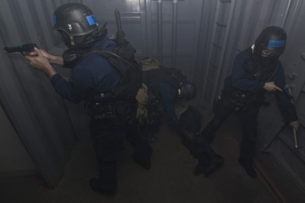 Eine Gruppe von Polizisten in Einsatzausrüstung, einer mit einer Waffe, neben einem Metallbehälter und einer Leiter in einem begrenzten Raum.