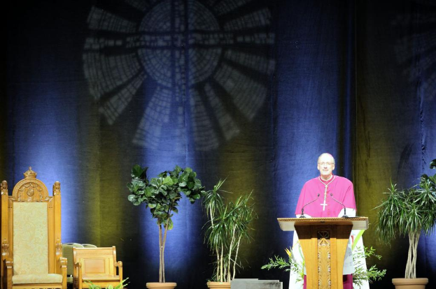 Ein Mann in einem rosafarbenen und weißen Kleid steht in einem Auditorium neben einem Rednerpult, mit Pflanzen und zwei Stühlen daneben und einer bunten Wand im Hintergrund.