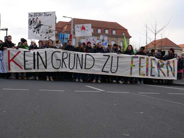 Eine Gruppe von Menschen steht auf der Straße und hält ein Transparent mit der Aufschrift "Kein Grund zu Feiern" in Protest, mit Straßenlaternen, Bäumen, Gebäuden und einem klaren blauen Himmel im Hintergrund.
