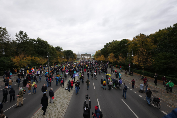 Eine große Gruppe von Menschen marschiert eine baumbestandene Straße in Berlin entlang und hält Kameras in der Hand, mit einem Gebäude und einem klaren Himmel im Hintergrund.
