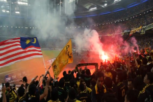 Große Zuschauermenge in einem Stadion mit Bengalos, Rauch und Schildern mit der Aufschrift "Malaysia vs Malaysia Live Streaming" bei einem Ereignis.