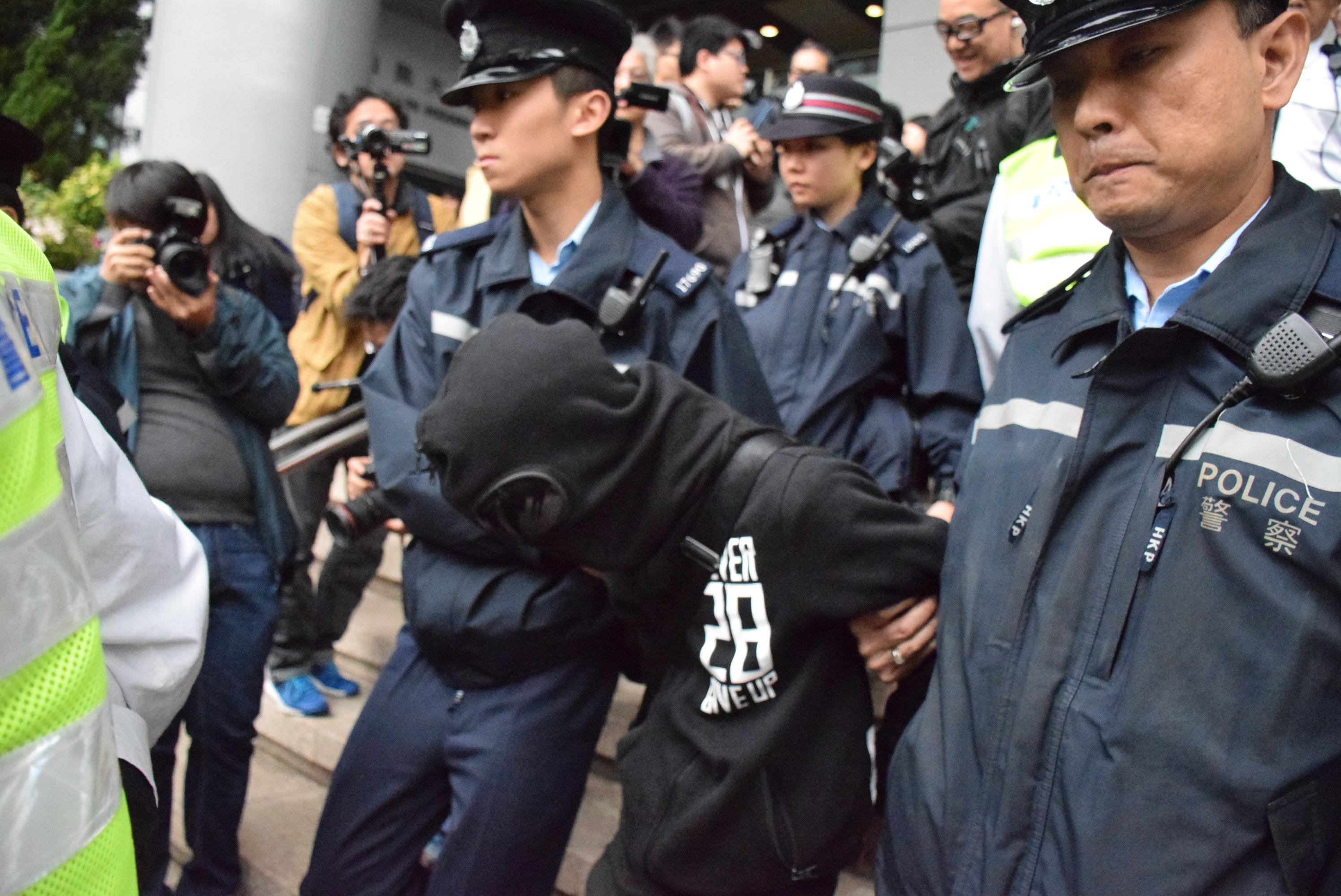 Eine Gruppe von Polizeibeamten, die vor einer Menschenmenge mit Säulen und Deckenbeleuchtung stehen, mit Bäumen links im Bild.