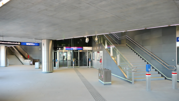 Ein leerer Bahnhof mit Treppen, Geländern, Mülltonnen auf dem Boden, Schildern mit Text, Deckenlampen und Glastüren im Hintergrund.