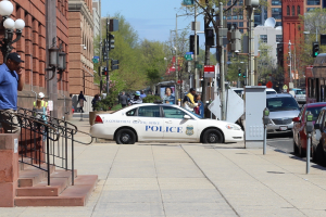 Eine belebte Stadtstraße mit einem geparkten Polizeiwagen, fahrende Fahrzeuge einschließlich eines Busses und eines Autos, Fußgänger auf dem Gehweg, Gebäude mit Fenstern und Geländern, Laternenpfähle, Bäume und einen klaren blauen Himmel.