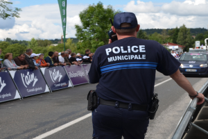 Ein Polizist in Uniform steht vor einer Menge und hält ein Funkgerät, mit Bannern, Fahrzeugen, Bäumen, Gebäuden und einem bewölkten Himmel im Hintergrund.