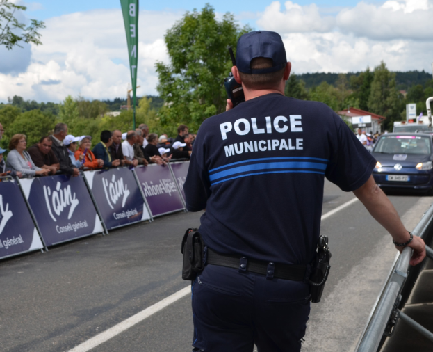 Ein Polizist in Uniform steht vor einer Menge und hält ein Funkgerät, mit Bannern, Fahrzeugen, Bäumen, Gebäuden und einem bewölkten Himmel im Hintergrund.