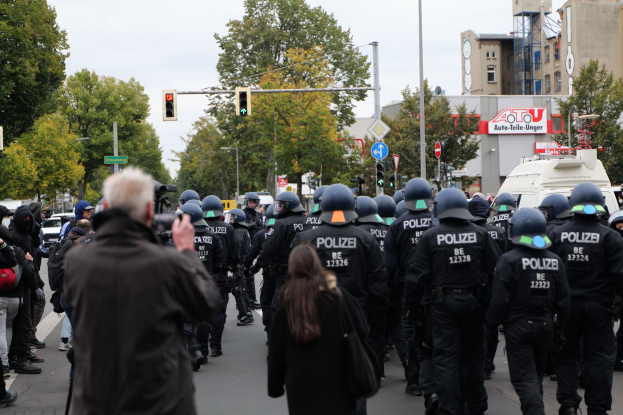 Eine Gruppe von Menschen geht eine Straße entlang, flankiert von Polizisten, einige tragen Helme und tragen Taschen oder Kameras, mit Fahrzeugen, Verkehrszeichen, Schildern, Bäumen, Gebäuden und einem klaren blauen Himmel im Hintergrund.