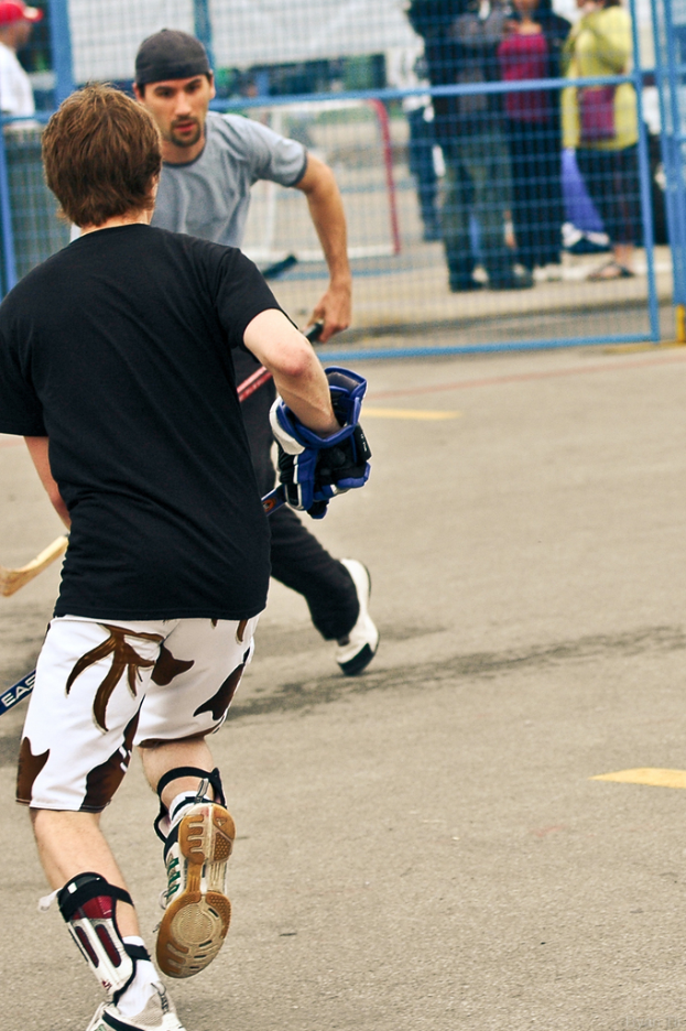Zwei Personen spielen Hockey auf einer Straße mit einem Zaun dahinter und einer Zuschauermenge im Hintergrund.