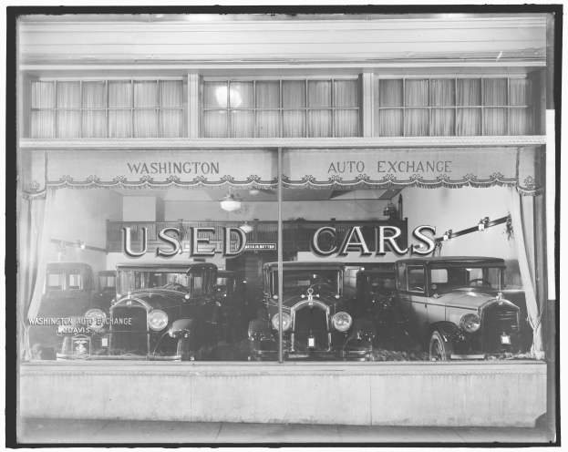 Schwarzes und weißes Foto eines Gebrauchtwagenhändlers in Washington, D.C., mit mehreren Autos durch die Glasscheibe sichtbar und Text an der Wand.