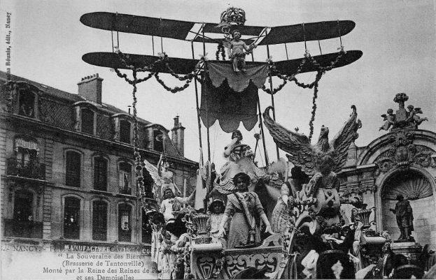 Ein Schwarz-Weiß-Foto eines Umzugswagens, der mit Statuen und einem Flugzeug geschmückt ist und eine Gruppe von Menschen trägt, mit einem Gebäude und Himmel im Hintergrund und Text unten, der darauf hinweist, dass es ein Foto vom Pariser Karneval ist.