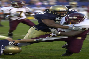 Zwei Fußballspieler in Sportkleidung und Helmen, einer hält den anderen auf dem Feld fest, mit zusätzlichen Spielern und Zuschauern im Hintergrund.