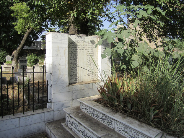 Ein Friedhof mit Treppe unten, Gras und Bäumen rechts, einem Gedenkstein in der Mitte, einem Geländer daneben, Gräbern dahinter und Bäumen und Gebäuden im Hintergrund.