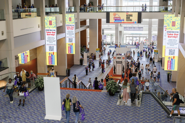 Eine große Menschenmenge, die durch ein großes Gebäude mit Bannern, Topfpflanzen, Rolltreppen, Geländern, Säulen, Texttafeln, Glastüren und anderen Gegenständen geht, einige tragen Taschen, wahrscheinlich beim Nationalen Buchfest.