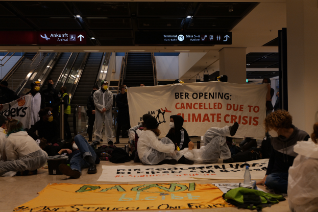 Eine Gruppe von Menschen sitzt auf dem Boden eines Flughafens und hält ein Schild in Händen, auf dem 'Berlin wegen Klimakrise abgesagt' steht, umgeben von verstreuten Taschen, Flaschen und anderen Gegenständen, und im Hintergrund sind Rolltreppen, Texttafeln und Deckenlampen zu sehen.