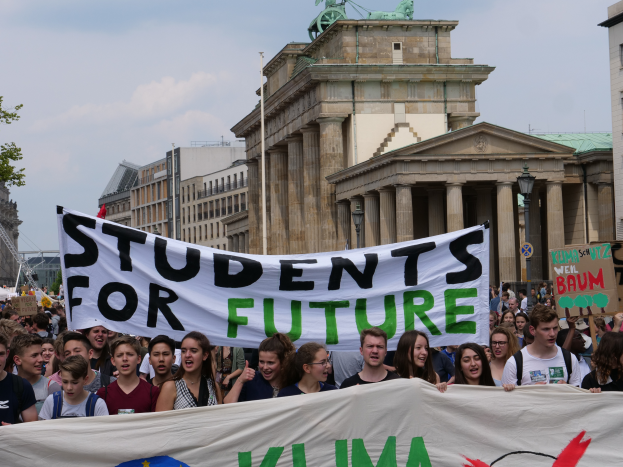 Eine Gruppe von Studenten marschiert in Berlin und hält ein buntes Banner mit der Aufschrift "Students for Future", mit Gebäuden, Bäumen und Himmel im Hintergrund.