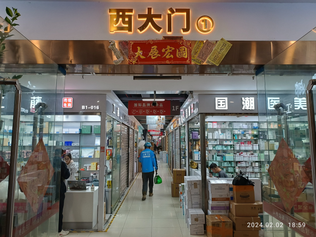Innenraum einer chinesischen Apotheke mit Glaswänden, Regalen voller Artikel, verstreute Kartons auf dem Boden, eine Pflanze links und ein Namensschild oben.