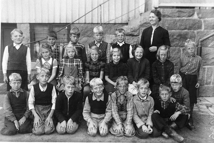 Schwarzes und weißes Foto einer Gruppe von Kindern, die vor einem Gebäude mit Treppe und Geländer posieren, mit der Beschriftung "1930er Schulkinder" unten.