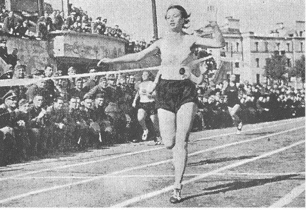 Schwarzes und weißes Foto einer Frau, die auf einer Laufbahn während des Frauen-100m-Finals bei den Olympischen Spielen 1956 läuft, mit Zuschauern und Gebäuden im Hintergrund.