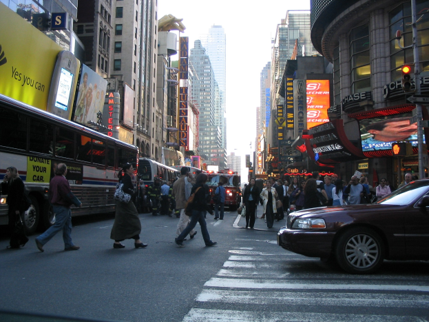 Eine belebte Stadtstraße mit Fahrzeugen, Fußgängern an einer Zebrastreifenfurt, Gebäuden, Beschilderung, Verkehrszeichen und einem blauen Himmel.