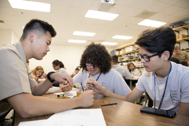 Three students at a table with papers, pens, a mobile phone, and other objects, working on a project in a classroom with racks, ceiling lights, and a projector.