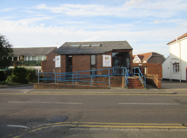 Ein Schulgebäude mit blauer Begrenzung, umgeben von Häusern, Treppen mit Begrenzungen, Informationsschildern, Pflanzen, Bäumen und einem bewölkten Himmel.