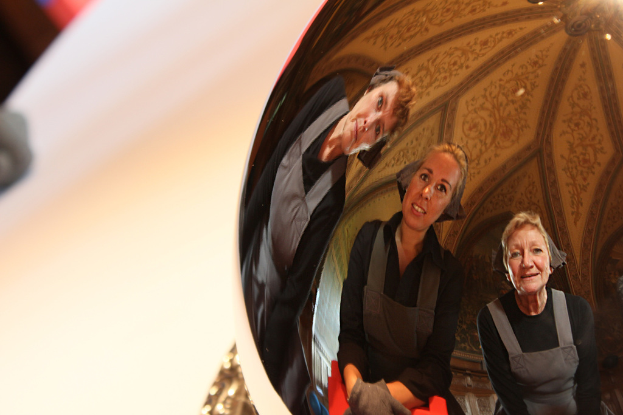 Ein reflektierender Ball auf einem weißen Tisch zeigt das Spiegelbild von drei lächelnden, leicht gebogenen Frauen mit einem Dach im Hintergrund.