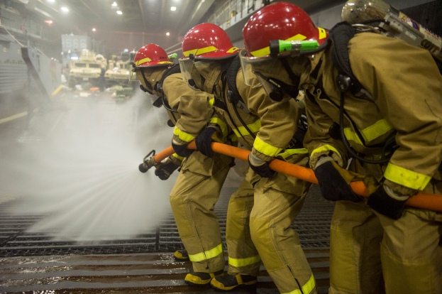 Feuerwehrleute in Helmen und Handschuhen sprühen Wasser aus Rohren auf ein Feuerwehrauto, mit verschiedenen Gegenständen und einem Boden im Hintergrund.
