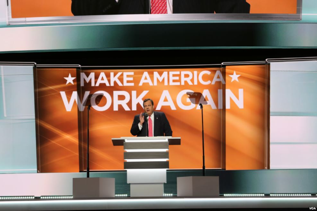 Ein Mann in einem schwarzen Blazer, weißem Hemd und roter Krawatte steht an einem Podium mit einem Mikrofon und spricht vor einem Bildschirm, auf dem 'Make America Work Again' steht, mit einem orangen Banner dahinter und zwei Personen in schwarzen Blazern und roten Krawatten oben auf dem Bild.