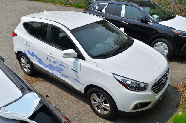 Drei Autos nebeneinander auf einem Parkplatz geparkt, wobei eines den Text 'wasserstoffbetriebenes Fahrzeug' anzeigt, ein Maschendrahtzaun im Hintergrund und Gras auf der rechten Seite.