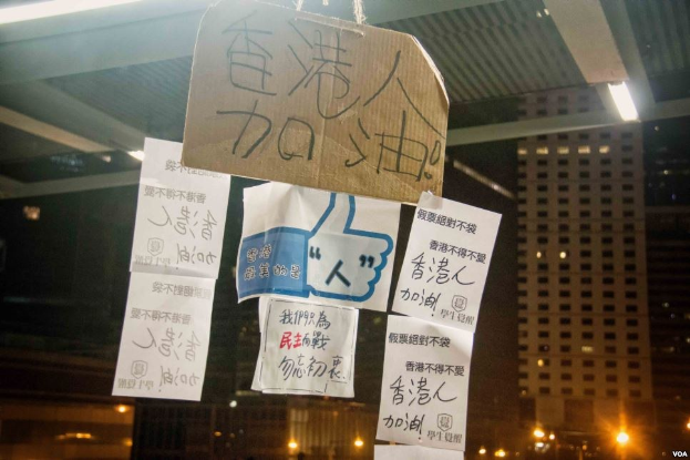 Gruppe von Menschen nachts vor einem Gebäude mit beleuchteten Protestschildern an der Decke.