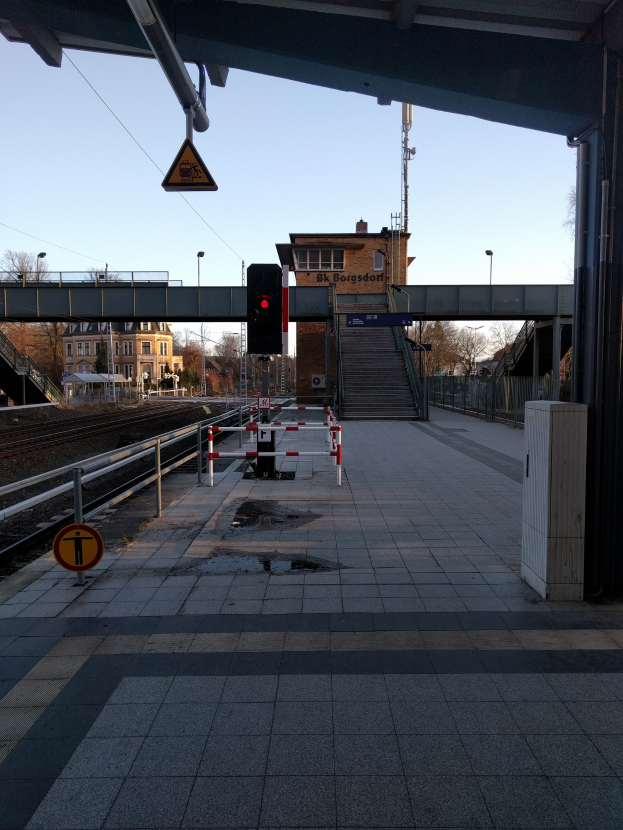 Ein belebter Bahnhof mit einem Zug auf den Gleisen, eine Plattform mit Geländern, ein Schild, eine Verkehrsampel an einem Mast, eine Brücke mit Treppen, Laternen, Bäume, Gebäude mit Fenstern, ein Turm und ein Himmel im Hintergrund.