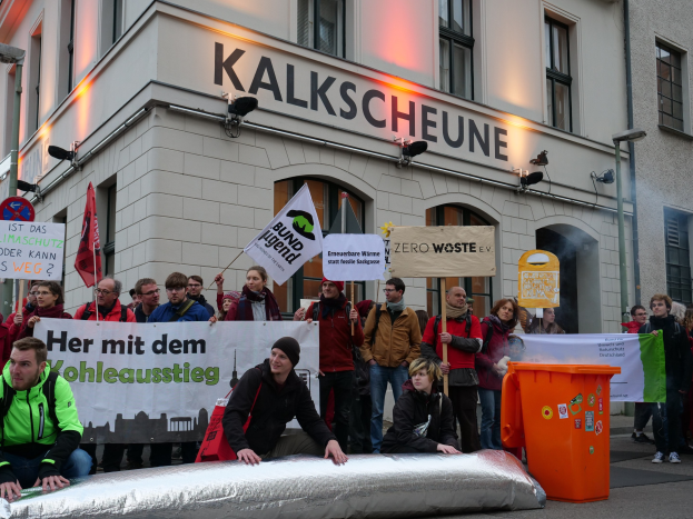 Eine Gruppe von Menschen steht vor einem Gebäude, hält Schilder und Plakate hoch, mit zwei Personen im Vordergrund und einem Müllcontainer auf der rechten Seite, während einer Demonstration in Deutschland.
